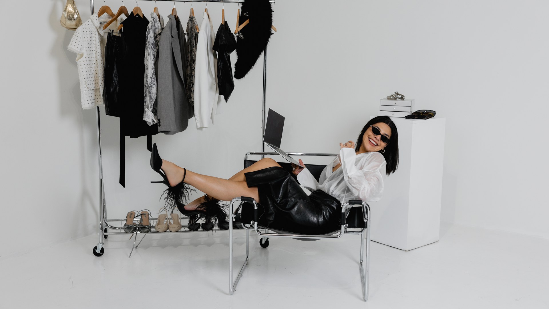 Camila Luna relaxing on a chair with her laptop beside a rack of styled pieces in her studio